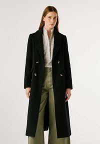 Cappotto nero doppiopetto con dettagli in hardware dorato, maniche lunghe e vestibilità sartoriale, abbinato a una blusa bianca a scacchi e pantaloni verdi.