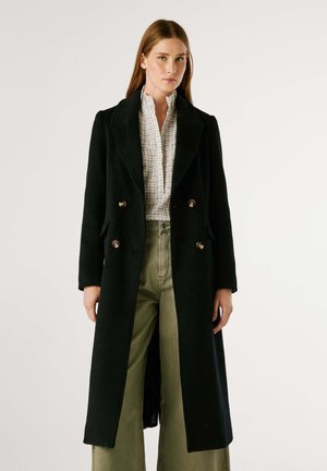 Cappotto nero doppiopetto con dettagli in hardware dorato, maniche lunghe e vestibilità sartoriale, abbinato a una blusa bianca a scacchi e pantaloni verdi.