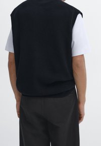 Ärmlös marinblå tröja med en texturerad stickning, bärs över en vit kortärmad skjorta, tillsammans med mörka byxor, sedd från baksidan.