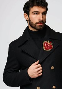 Schwarzer Wollmantel mit doppelreihigem Schnitt, verziert mit goldenen Knöpfen und einer großen roten brosche in Form eines juwelenbesetzten Herzens am linken Revers.