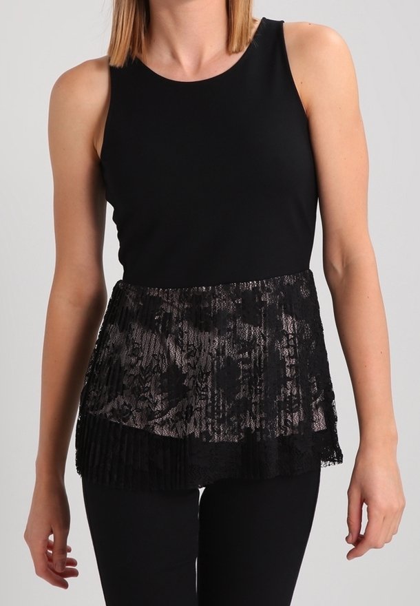 Femme portant un top noir sans manches avec un détail basque en dentelle et un pantalon noir sur un fond clair uni.