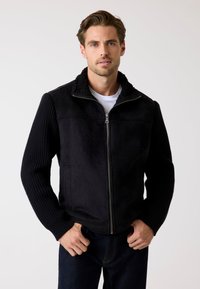 Schwarze Jacke mit Reißverschluss vorne, Fleece-Kragen und strukturierten Strickärmeln. Wildleder-Körper mit zwei Seitentaschen, kombiniert mit einem weißen Hemd.