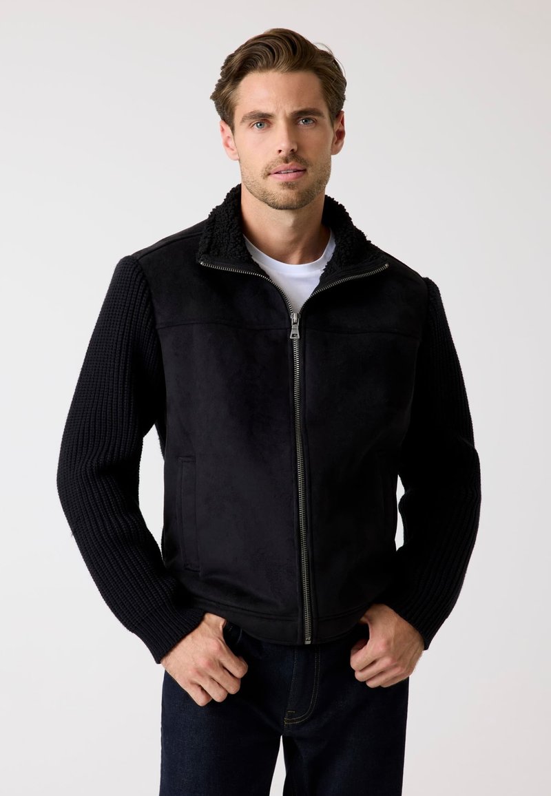 Schwarze Jacke mit Reißverschluss vorne, Fleece-Kragen und strukturierten Strickärmeln. Wildleder-Körper mit zwei Seitentaschen, kombiniert mit einem weißen Hemd.