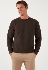 Hanorac maro cu mâneci lungi și decolteu rotund, realizat dintr-un material moale, având o textură subtilă și manșete ajustate. Asortat cu pantaloni bej.