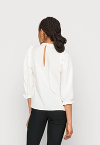 Blouse blanche avec des épaules froncées, un dos en forme de clé et des poignets plissés. Texture lisse, associée à un pantalon noir ajusté.