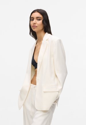 Ženska z dolgimi temnimi lasmi, oblečena v ohlapen bel blazer, bele hlače in črno kratko majico, na preprostem svetlem ozadju.