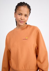 Orange sweatshirt med rund halsudskæring og ribbede manchetter. Har et lille Carhartt-logo i gul og burgunder på brystet.