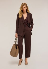 Donna bionda in un completo scuro marrone su misura e tacchi beige tiene una grande borsa beige, in piedi contro uno sfondo chiaro uniforme.