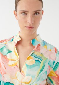 Blouse à col avec un motif floral dans des couleurs pastel, y compris le rose, le jaune et le bleu, dotée d'une texture légère et d'une coupe décontractée.