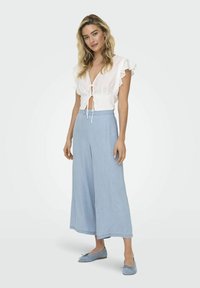 Blusa blanca de manga corta con cierre de lazo y mangas con volantes, combinada con pantalones de pierna ancha azul claro y bailarinas a juego.