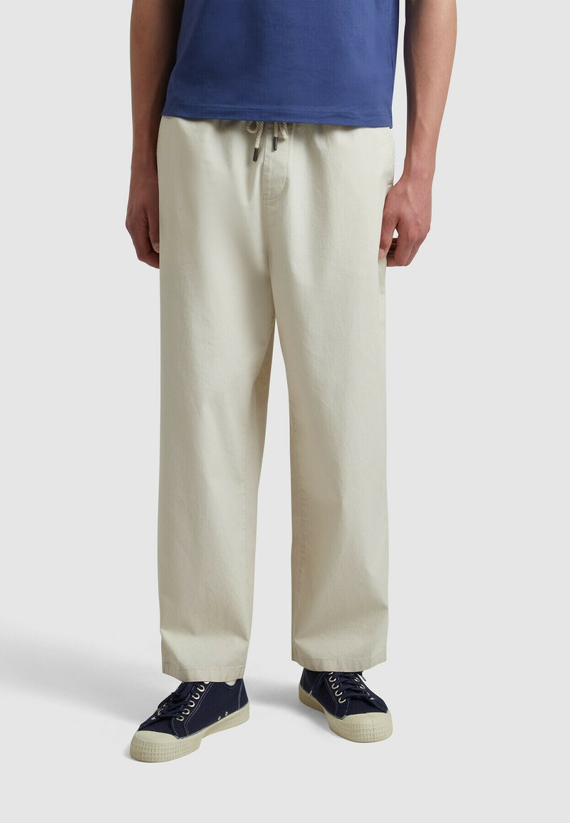 Helle beigefarbene Hose mit relaxed Fit, ausgestattet mit einem Kordelzugbund, umgeschlagenen Säumen und kombiniert mit navyfarbenen Turnschuhen. Glatte Baumwolltextur.