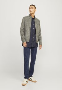 Gray suede bomber jacket with a zip closure, navy button-up shirt, blue jeans, and cream sneakers featuring green accents. Flat design, casual fit.