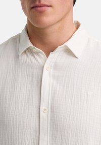 Witte blouse gemaakt van gestructureerde stof, met een knoopsluiting aan de voorkant en een klein geborduurd logo op de borst.