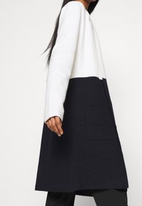 Cappotto color block bianco e blu navy con maniche lunghe, realizzato in tessuto morbido, dotato di tasca a toppa sul davanti e vestibilità comoda.