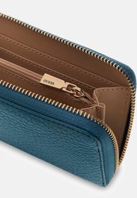 Portafoglio blu con texture e cerniera dorata, con interno marrone e scomparto con cerniera. Nome del marchio visibile sull'hardware.