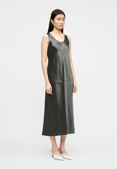 Γυναίκα με μακριά μαύρα μαλλιά, φορόντας ένα αμάνικο, γυαλιστερό, σκούρο γκρίζο midi φόρεμα και άσπρα μυτερά παπούτσια με τακούνι, στέκεται σε λευκό φόντο.