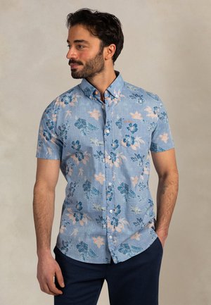 Man staat met handen in de zakken, draagt een lichtblauw overhemd met korte mouwen en een roze en blauwe bloemenprint.