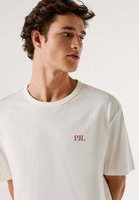 T-shirt bianca in cotone con scollo rotondo, caratterizzata da un piccolo ricamo rosso "PJL" sul lato sinistro. Maniche corte e tessuto liscio.