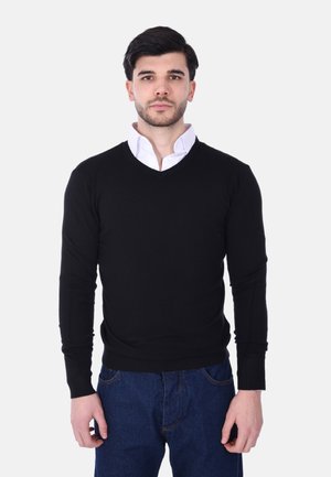 Maglione nero a collo a V con un design aderente, realizzato in materiale morbido, con polsini e orlo a coste. Indossato sopra una camicia bianca con colletto.