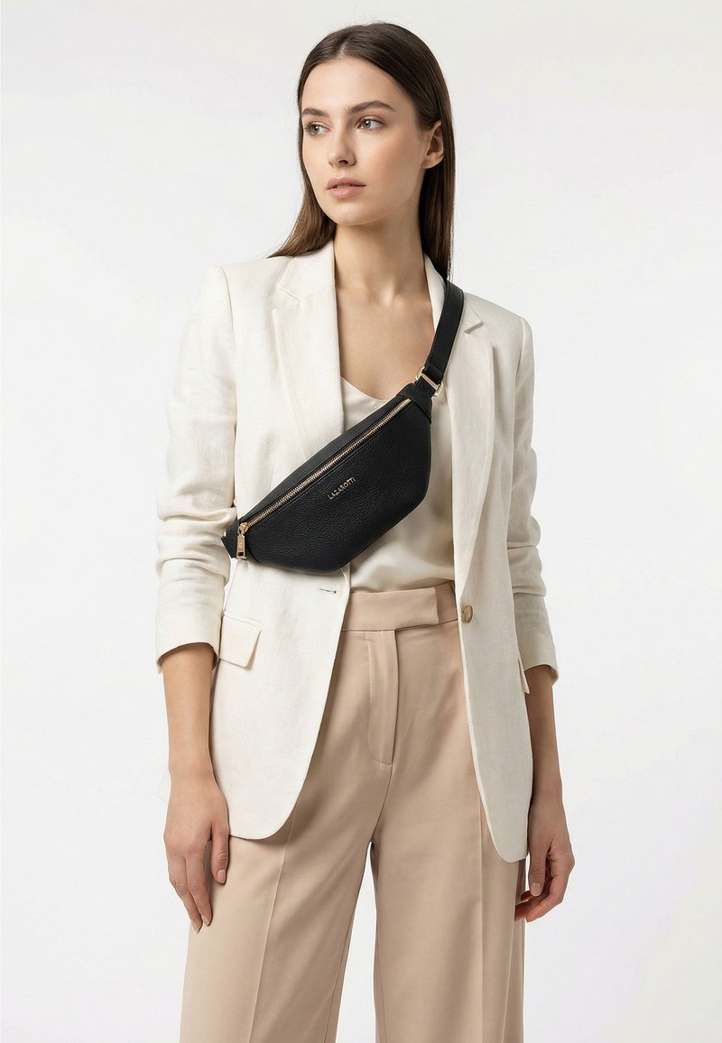 Donna che indossa pantaloni beige e blazer bianco con borsa a tracolla in pelle nera, guarda verso sinistra su sfondo bianco.