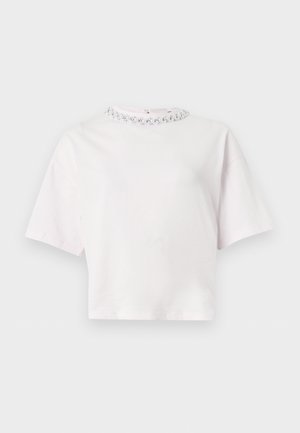 Kvinde iført hvid top med korte ærmer og lyserøde shorts, stående med hænderne på hoften, brune loafers med frynser, neutral baggrund.