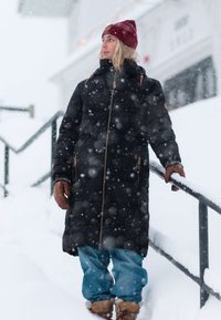 Person wearing a long black coat, red beanie, and brown gloves standing on snowy steps, holding a snow-covered railing during snowfall.