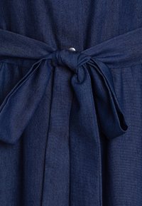 Granatowa sukienka dżinsowa z zawiązywanym paskiem w talii, detalami w postaci guzików i fakturowanym materiałem w dopasowanym kroju. Materiał jest gładki z delikatnym połyskiem.