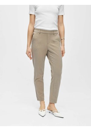 Femme portant un pantalon beige ajusté coupé à la cheville, un haut blanc à manches courtes, un bracelet argenté et des chaussures blanches à talons bas pointues.