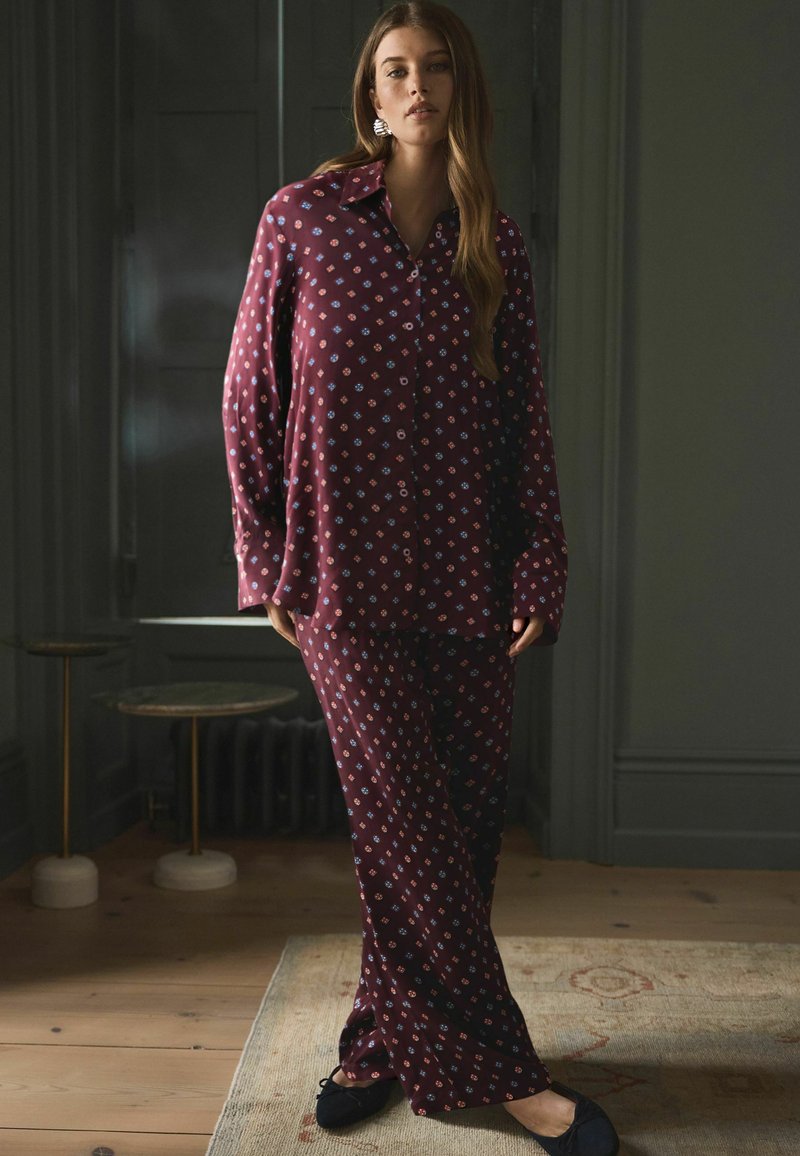 Mörkröd pyjamajacka med mönstrad design; långärmad skjorta och vida byxor. Slät tyg och lös passform för komfort. Svarta slip-on skor.