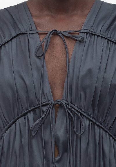 Κοντινό πλάνο σε στήθος με σκούρο δέρμα, με γκρι φόρεμα που έχει δύο δεμένους φιόγκους στον λαιμό και στη μέση του κορμού, με λεπτομέρειες από μαζεμένο ύφασμα.