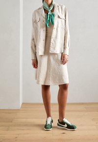 Chaqueta de lino crema con dos bolsillos frontales sobre un vestido a juego. Una bufanda verde acentúa el conjunto. Zapatillas retro verdes y blancas completan el look.