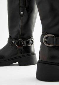 Schwarze Lederstiefel bis zum Knie mit runder Zehenform, matter Oberfläche, Nähdetails und einem dekorativen Schnallenakzent am Knöchel.