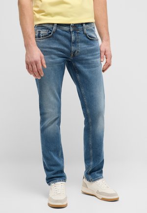 Mann trägt blaue Slim-Fit-Jeans, gelbes Hemd und weiße Sneakers, steht vor einem einfarbigen hellgrauen Hintergrund.