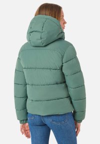 Padded jacket w stonowanym zielonym kolorze, z kapturem, poziomym przeszyciem i elastycznymi mankietami. Połączony z niebieskimi dżinsami.