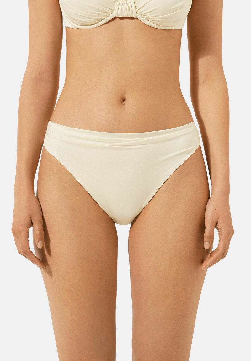 Bas de bikini couleur crème avec une texture lisse, couverture moyenne et une large taille. Design simple, sans motifs ni accents visibles.