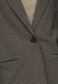 Brown and black checkered blazer featuring a notched collar, single button closure, and two front pockets. Textured fabric underlay visible.