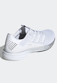 Zapatilla atlética blanca con una parte superior de malla transpirable, perforaciones sutiles y una suela texturizada. Presenta tres rayas diagonales y un acento plateado.