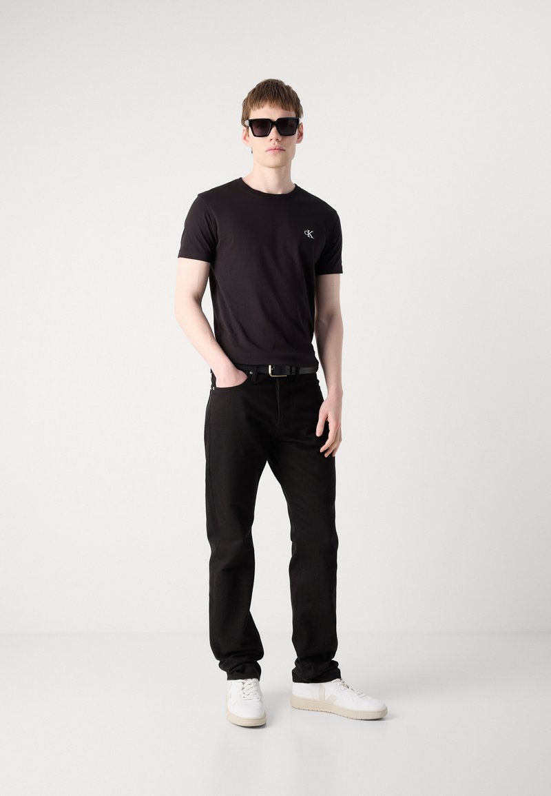 Hombre con camiseta negra de Calvin Klein, pantalones negros, zapatillas blancas y gafas de sol negras, de pie con una mano en el bolsillo.