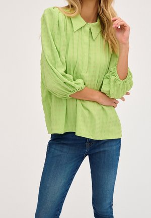 Femme portant un chemisier vert clair à manches longues avec col et un jean bleu, bras croisés et une main près du visage.