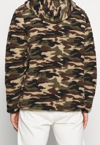 Mikina s kapucí z fleecu v maskáčovém vzoru hnědé, zelené a černé barvy s jemnou texturou, opatřená předním zipem a bočními kapsami.