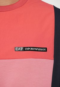 T-shirt z krótkim rękawem z kolorową blokadą w odcieniach koralowym i różowym, ozdobiony czarną metką z logo marki umiejscowioną na dole.