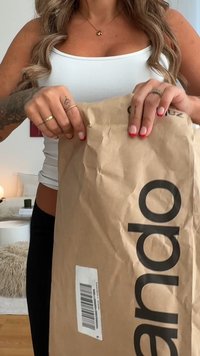 Bruine papieren verpakking met vetgedrukte zwarte letters, die met beide handen wordt geopend. Rimpelige textuur en een zichtbare barcode aan de zijkant.