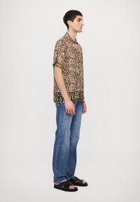 Camisa de manga corta con estampado de leopardo y dobladillo negro, con texto blanco, combinada con jeans de mezclilla azul claro y sandalias negras. Vista lateral.