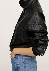 Veste en cuir synthétique noir avec un col montant, des poches latérales et des poignets à boutons, portée avec un jean en denim bleu clair. Texture lisse, design structuré.