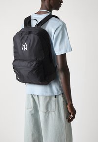 Zaino in nylon nero con un grande logo bianco, tasca frontale con zip e spallacci regolabili, indossato sopra una camicia azzurro chiaro e jeans chiari.