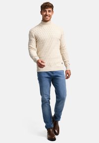 Suéter de punto trenzado crema con cuello alto y patrón texturizado, combinado con jeans azul claro y botas de tobillo marrón oscuro.