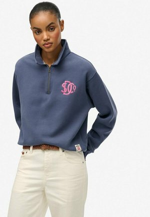 Marineblå fleece-pullover med et lyserødt grafisk logo, halv lynlås krave og lange ærmer; båret med cremede bukser og et fletbælte.