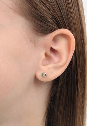 Primo piano di un orecchio di donna con capelli castani lisci che indossa un piccolo orecchino a forma di fiore in oro con gemme verdi.