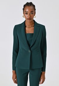 Blazer su misura verde scuro con rever a becco, chiusura con un bottone e due tasche frontali. Tessuto liscio con una silhouette aderente.
