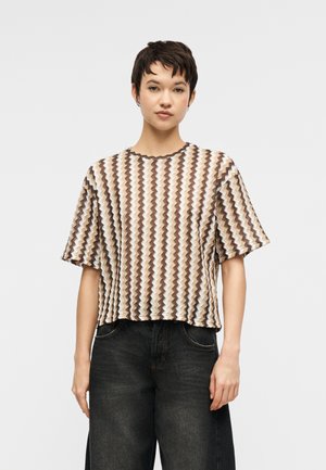 Kobieta z krótkimi ciemnymi włosami, ubrana w luźną bluzkę na krótki rękaw o zygzakowatym wzorze w kolorach brązu, beżu i bieli, w połączeniu z czarnymi jeansami z wysokim stanem.